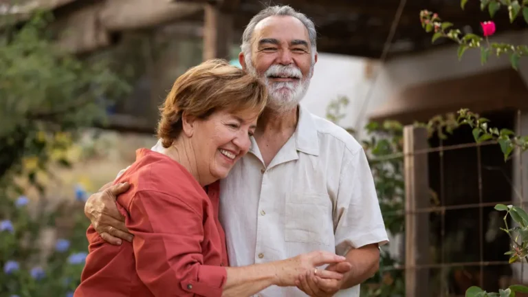Older couple hugging in a garden near a countryside home, sharing a calm, happy moment.