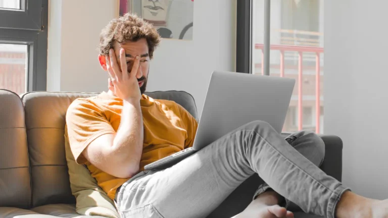 Tired man on a sofa with a laptop, covering his face with one hand in bright window light.