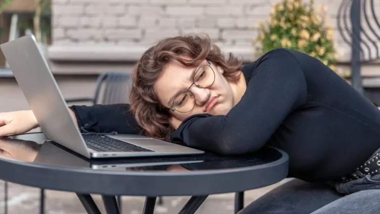 Exhausted woman with glasses resting head on arms next to laptop showing December burnout and year-end fatigue symptoms