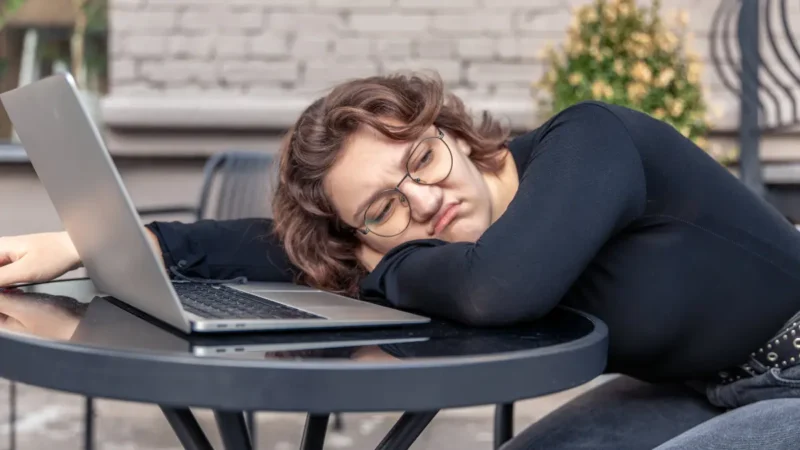 Exhausted woman with glasses resting head on arms next to laptop showing December burnout and year-end fatigue symptoms