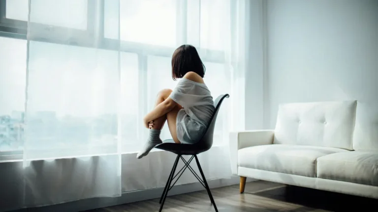Young woman in white t-shirt sitting alone on chair by window, gazing outside in minimal living room, feeling isolated or contemplative