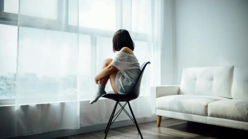 Young woman in white t-shirt sitting alone on chair by window, gazing outside in minimal living room, feeling isolated or contemplative