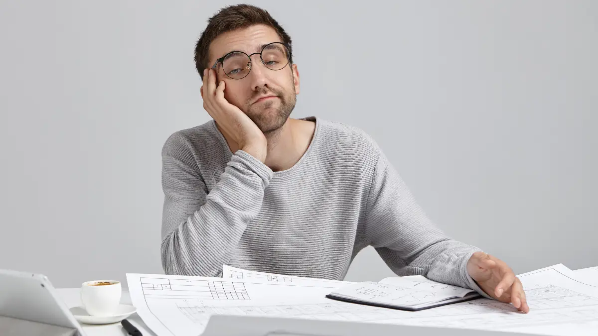 Stressed male professional with glasses looking exhausted at desk with blueprints displaying year-end burnout and fatigue