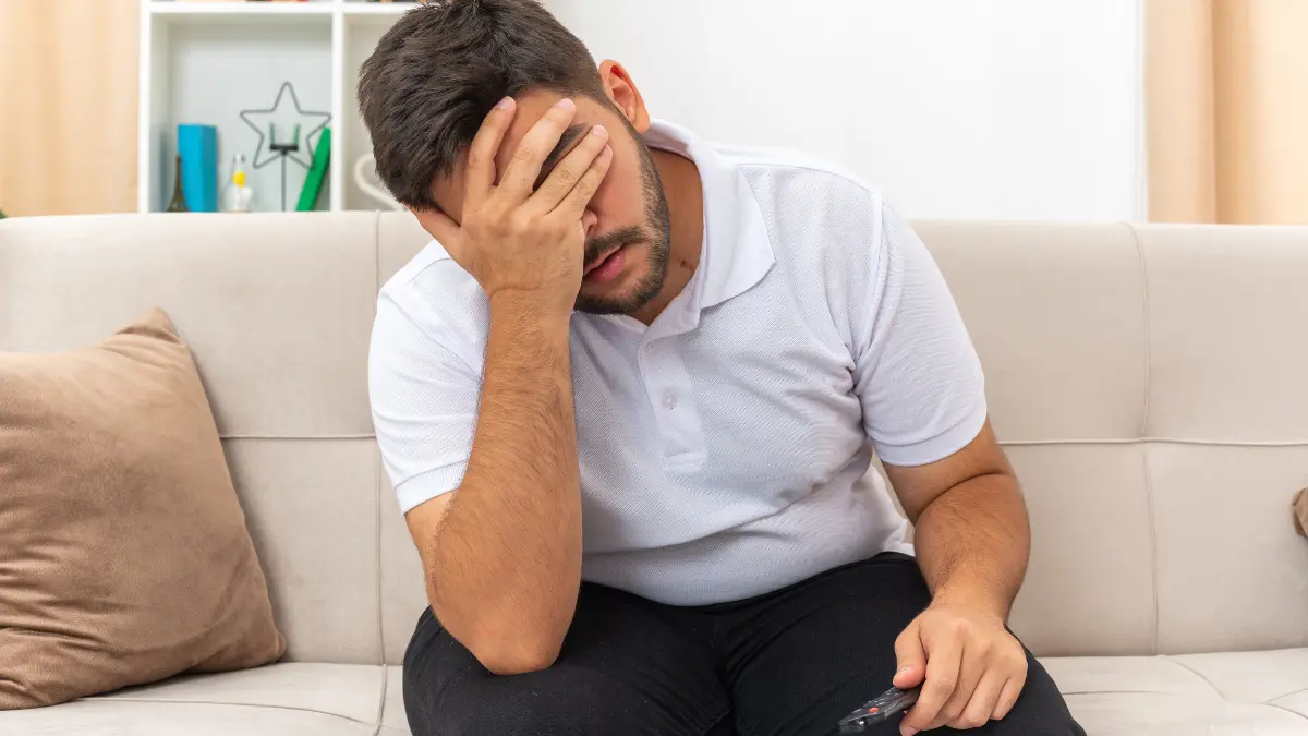 Man showing clinical end of year burnout symptoms covering face with hands experiencing December anxiety and emotional exhaustion on couch