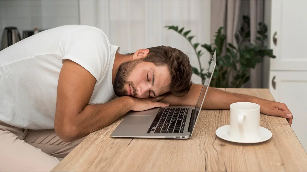 Man asleep on his arm at a table beside an open laptop and a cup of coffee.