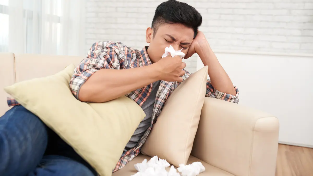 Young Asian man in plaid shirt holding tissue to nose suffering from post-Christmas cold flu and respiratory infection symptoms at home December 2025