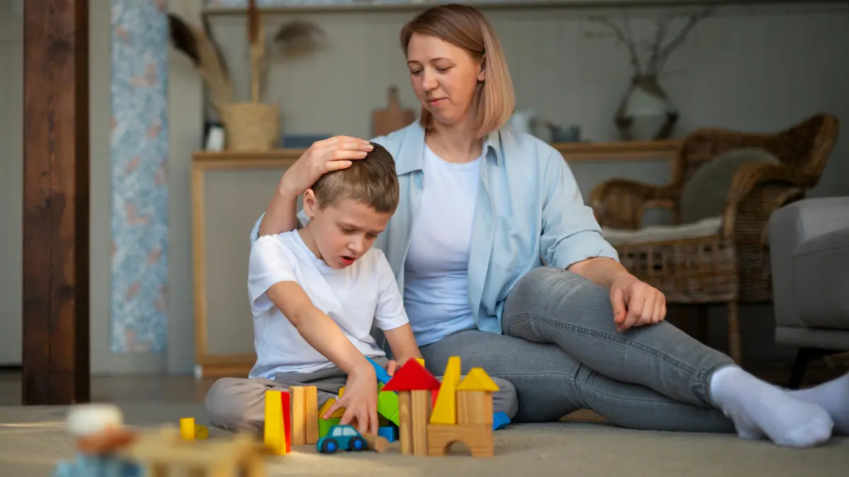A parent sits on the floor with a child, gently resting a hand on the child’s head as they play with colorful wooden blocks at home.