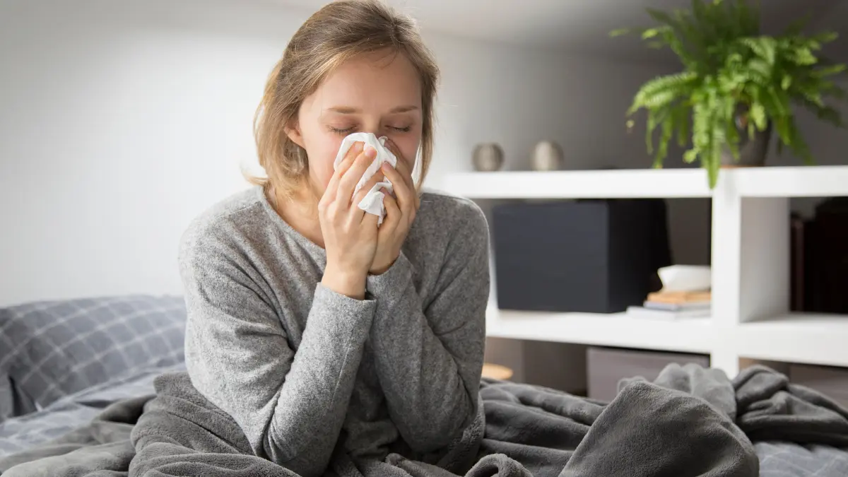 Woman with severe flu symptoms sitting in bed blowing nose with tissue during 2025 influenza season
