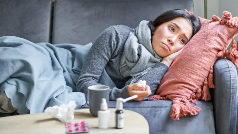 Young woman lying sick on gray couch wrapped in blue blanket with medications on table suffering from post-Christmas flu and respiratory virus after holiday gathering December 2025