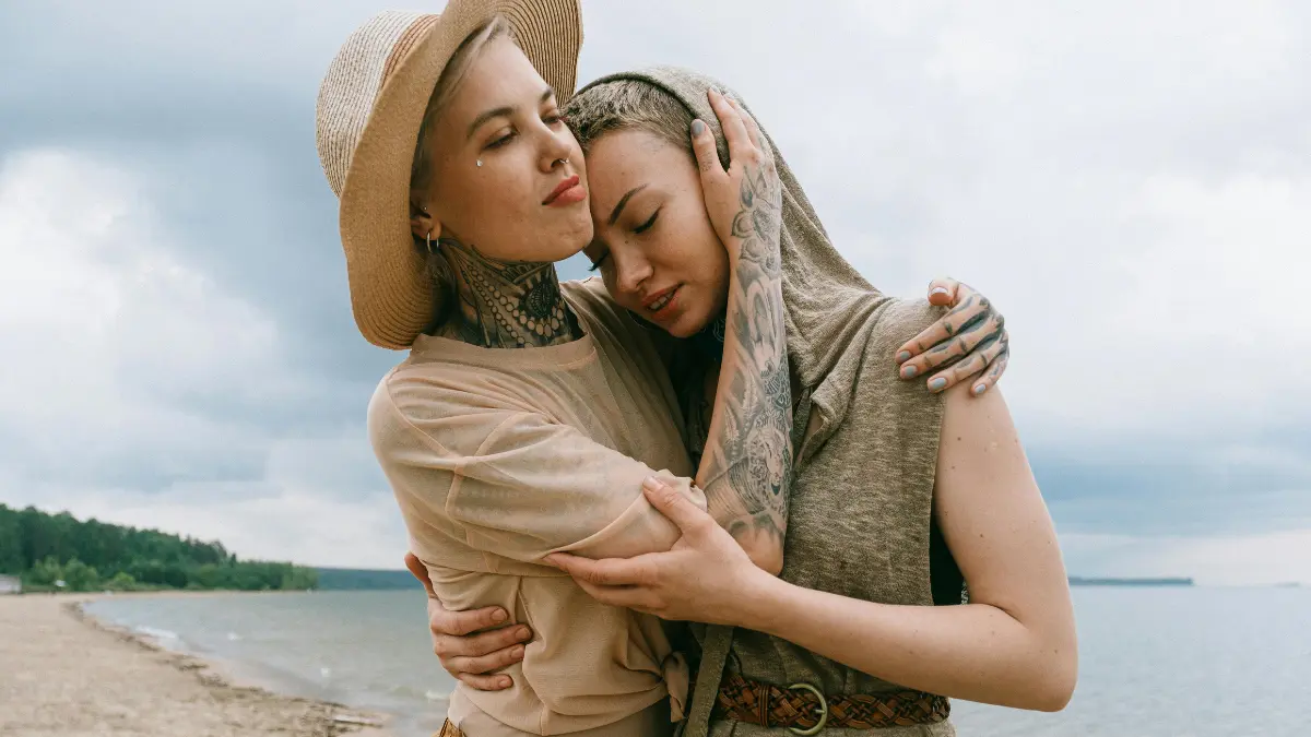 Two adults embrace on a quiet beach, one comforting the other.