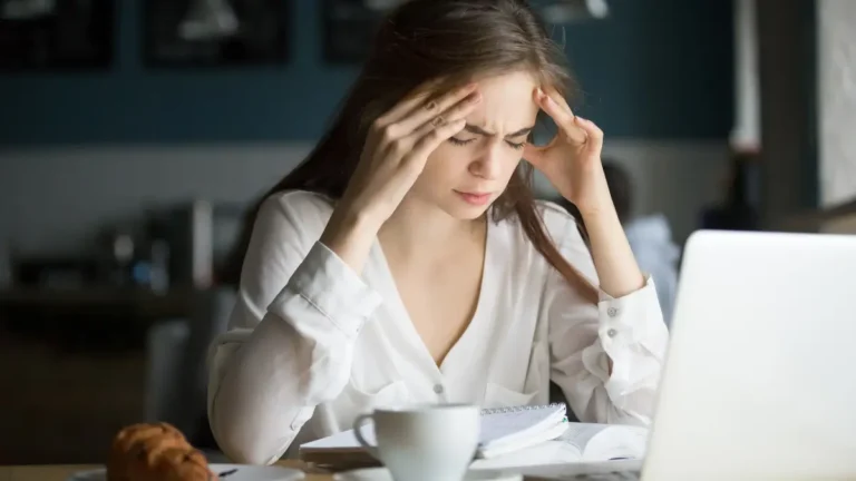 Woman holding her temples while feeling a wave of quiet fatigue at a café table
